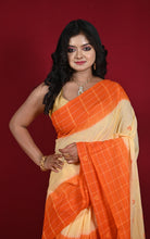 A woman modeling a traditional Indian saree with a vibrant orange and pale yellow color-blocked design, set against a solid dark red studio background.
