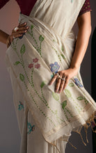 Close-up detail of hand-woven purple floral vines, green leaves, and bird motifs on off-white Jamdani saree fabric.
